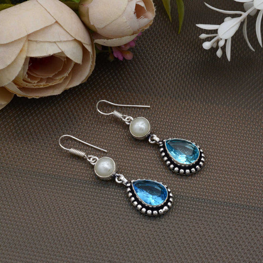 Pair of silver earrings with blue teardrop and pearl accents on a brown surface with flowers.