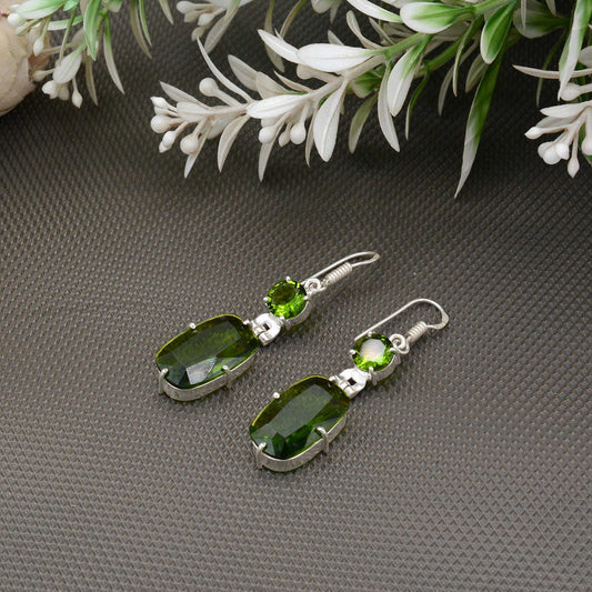 Green gemstone earrings on a gray surface with green leaves in the background